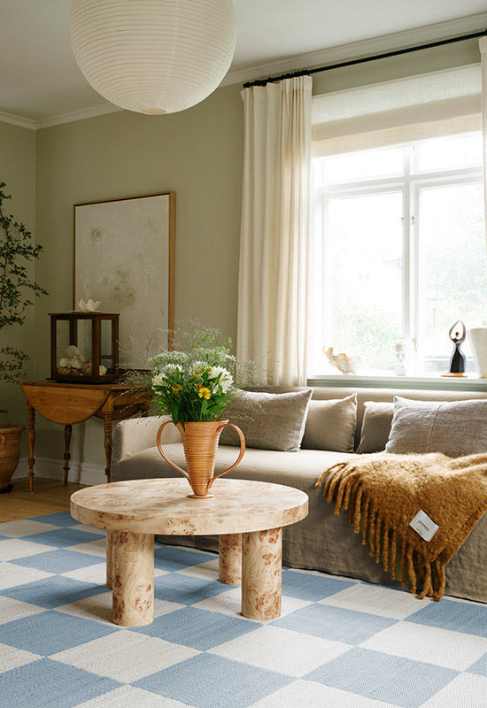 Layered Chess Cornflower Blue rug styled with pale wood furniture and soft grey upholstery in a light-filled living room